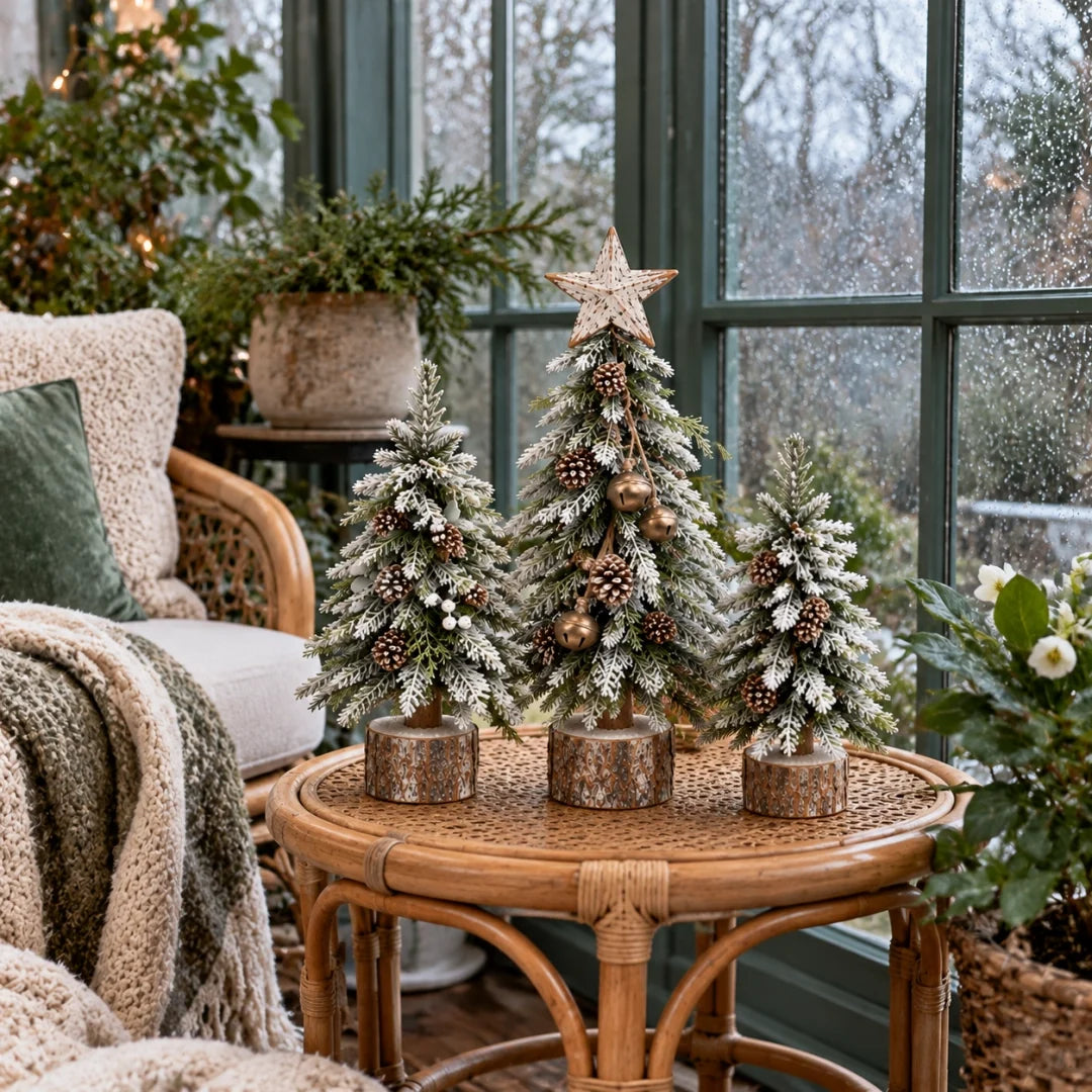 A three-piece snowy Christmas tree arrangement by a sunroom window